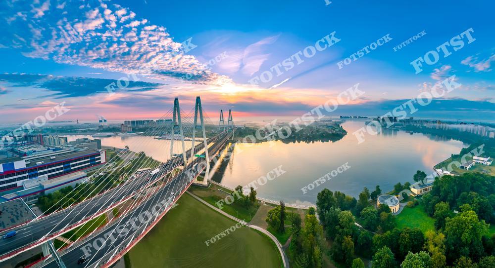 На фотографии запечатлен Большой Обуховский мост, один из самых известных вантовых мостов Санкт-Петербурга. Рассветное небо переливается оттенками розового и голубого, отражаясь в спокойной воде реки Невы. Вокруг моста расположены жилые кварталы и зелёные зоны, подчеркивающие современный облик города. Панорамный вид позволяет насладиться гармонией урбанистического пейзажа и природы.